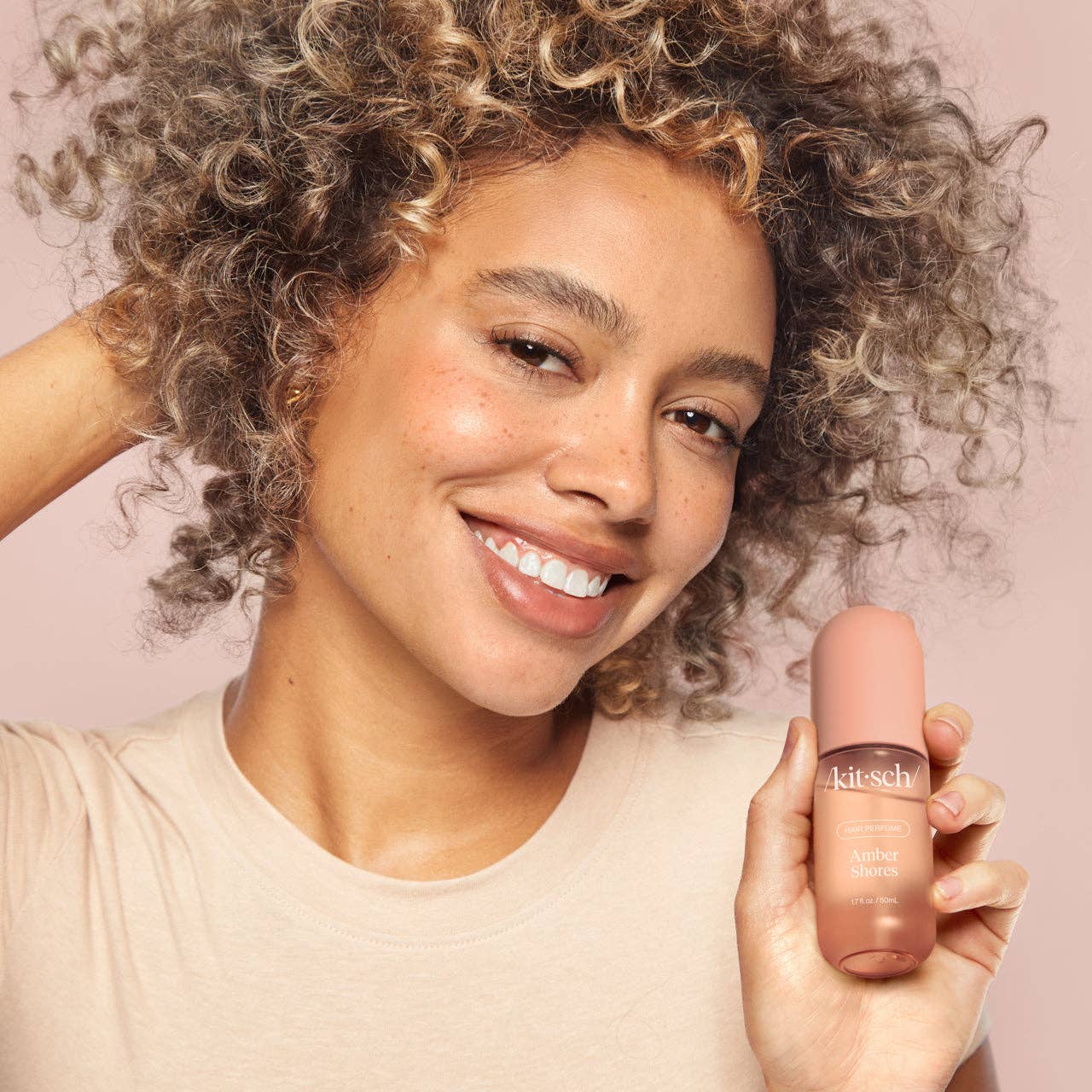 Smiling woman holds Amber Shores Hair Perfume bottle, woody aquatic scent of sea salt, amber and driftwood.