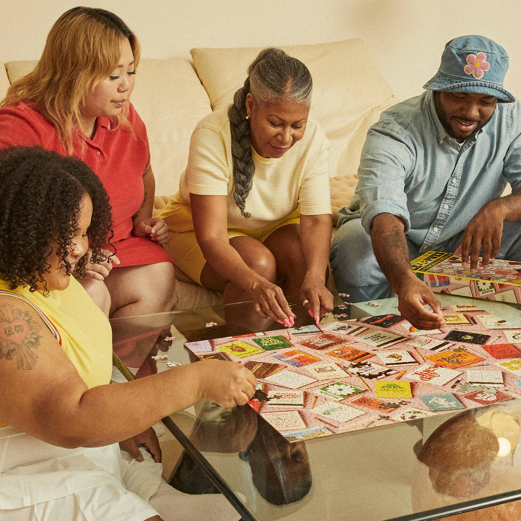 Four friends assemble Match Made in Heaven 1000 Piece Puzzle featuring vintage matchbook covers on a glass table.